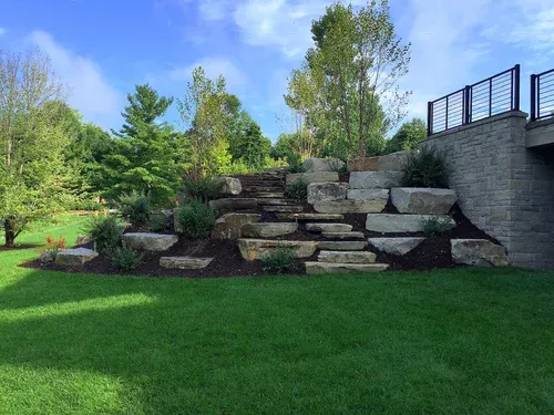 Stone retaining wall and steps in a grassy backyard garden under a blue sky.