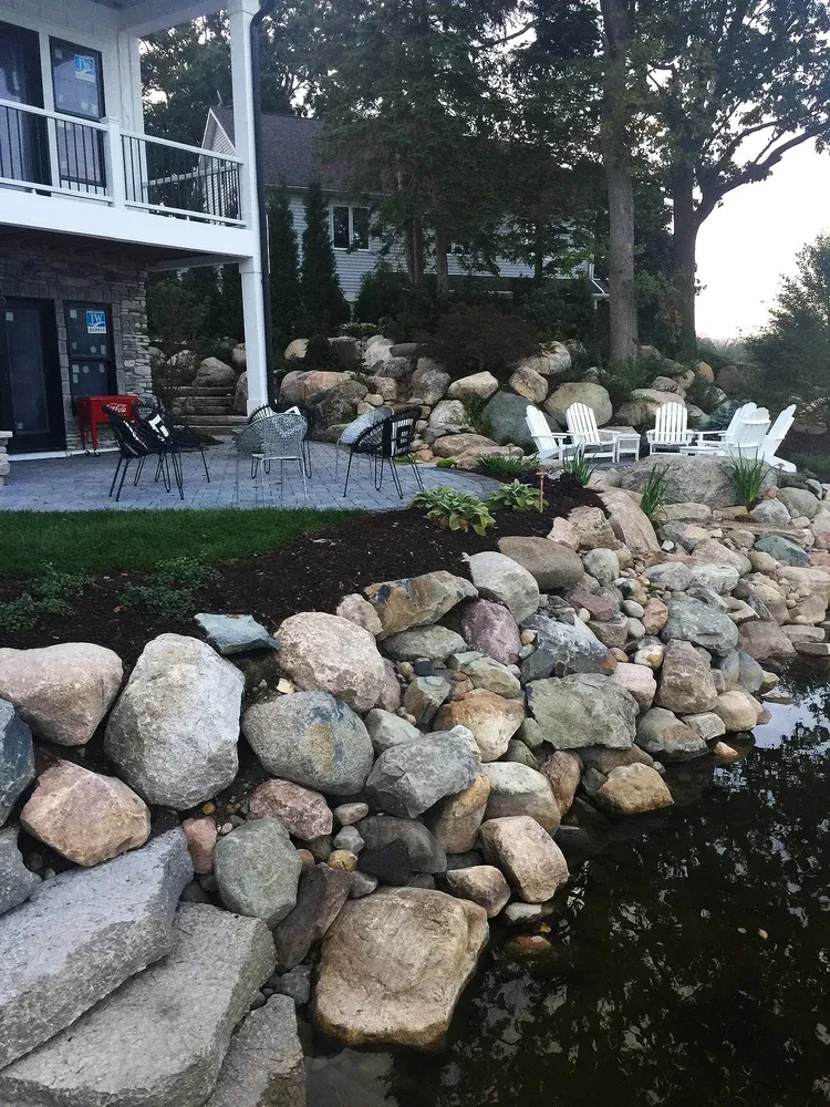 Rock retaining wall beside a patio and house, with outdoor chairs and trees in a landscaped yard.