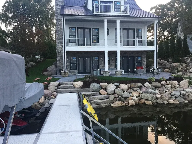 Waterfront house with white porch, stone retaining wall, dock, and calm water in the foreground
