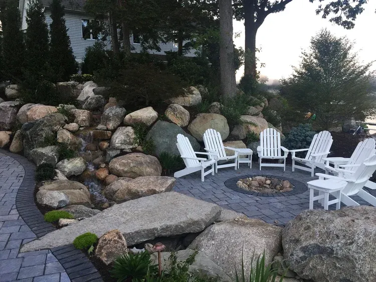 Stone patio with white chairs around a fire pit beside a landscaped rock garden