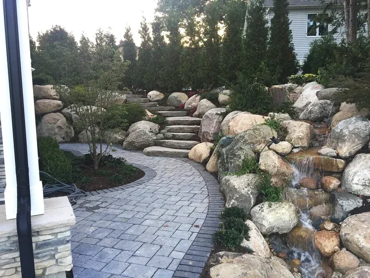 Curved stone garden walkway beside stacked rocks, shrubs, and steps in a landscaped yard