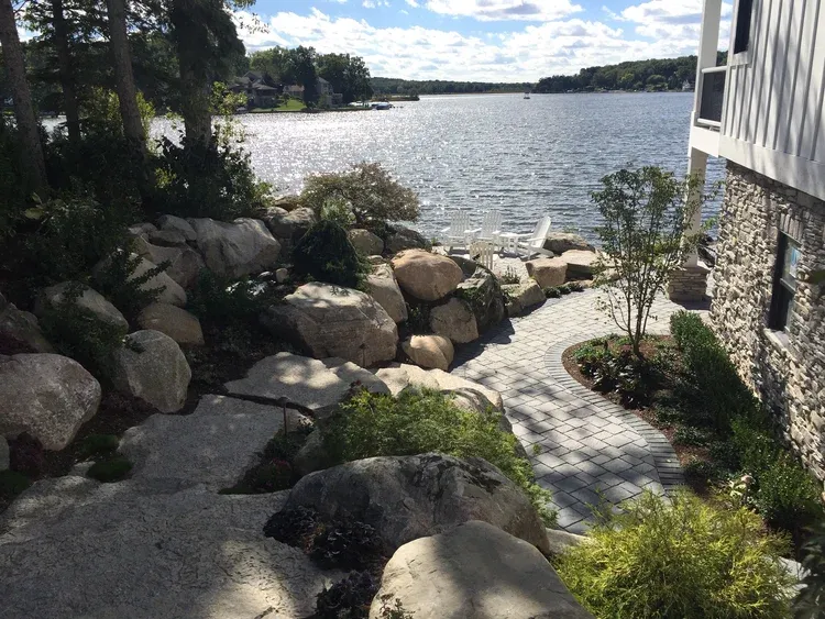 Rocky lakeside garden path beside a house, with water and boats in the distance.