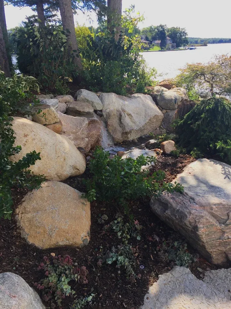 Rocky garden by a lakeside, with large boulders, green shrubs, and water in the background