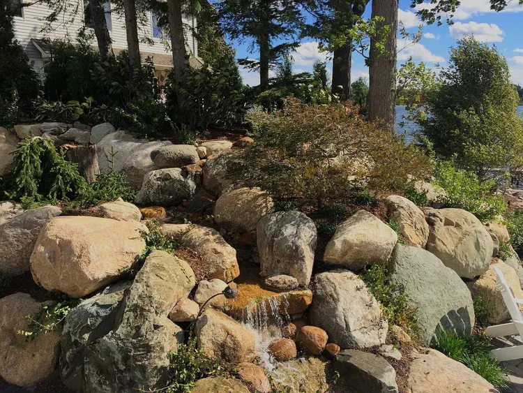 Rock garden waterfall with stacked boulders, flowing water, and greenery in a sunny landscaped yard
