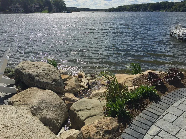 Rocky lakeshore with calm water, large stones, plants, and a curved brick path under a sunny sky