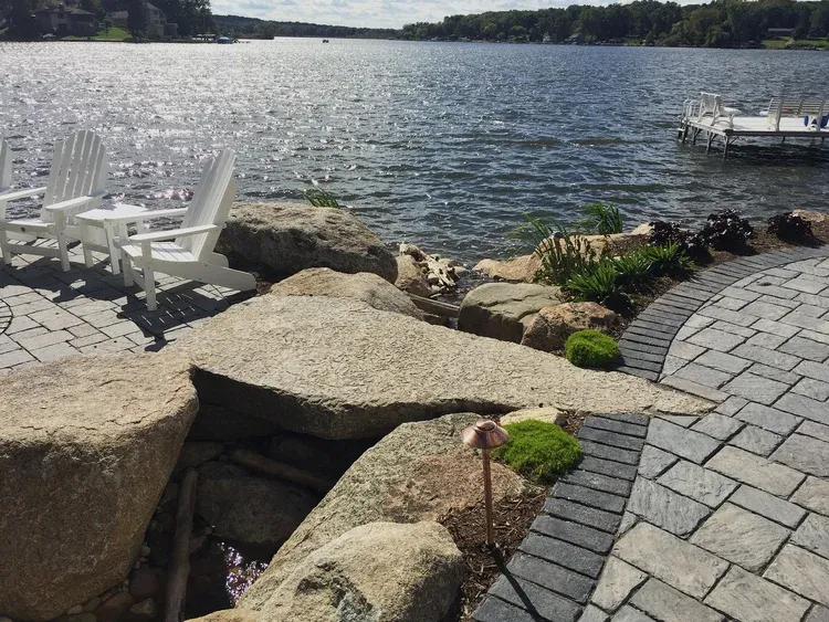 Stone patio overlooking a lake, with white chairs on the left and a dock on the right.
