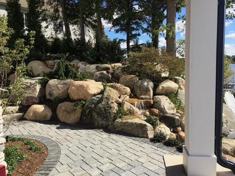 Stone retaining wall with shrubs beside a curved brick patio and white column in a sunny garden.