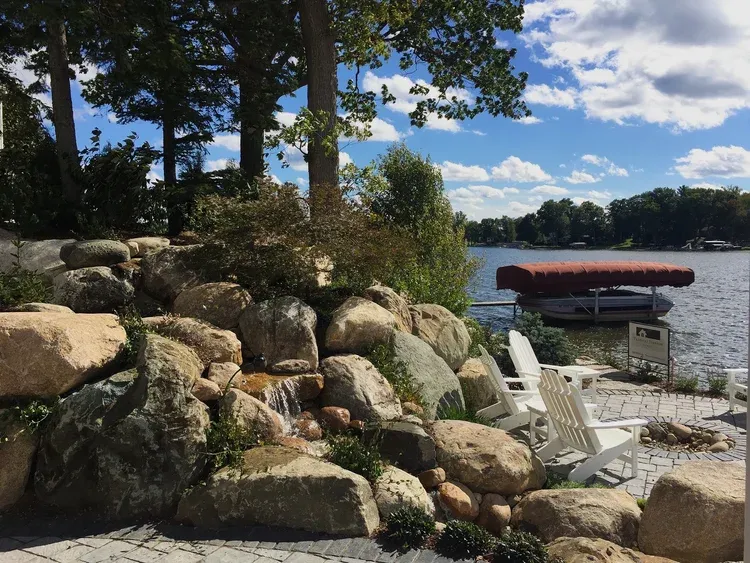 Rocky lakeside with white chairs, stacked canoes, and trees under a partly cloudy sky