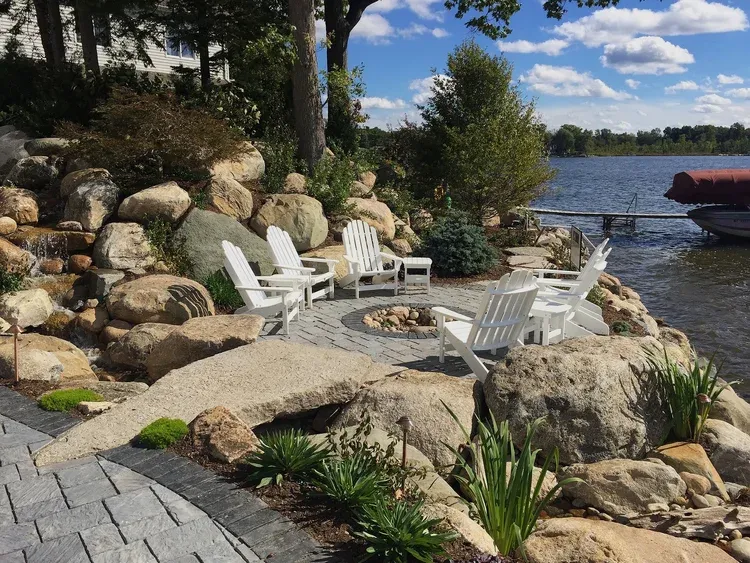 Stone fire pit with white chairs on a lakeside patio, beside rocks, plants, and a dock with a boat.