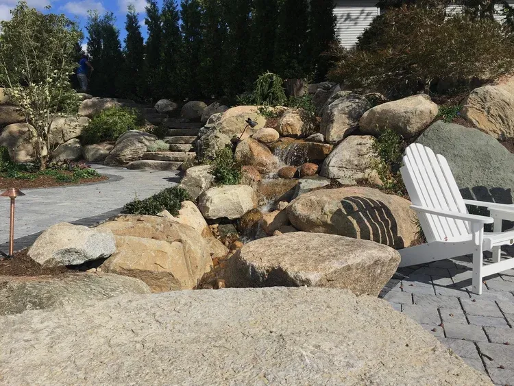 Rock garden landscape with boulders, shrubs, and white patio chairs on a stone patio