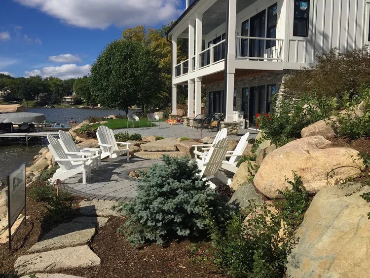 Lakeside patio with white chairs, stone path, and a modern house beside landscaped rocks and greenery