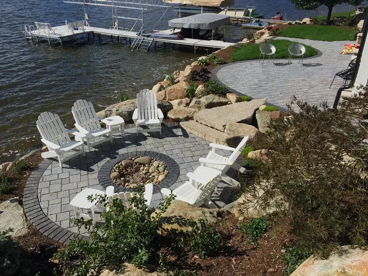Lakeside fire pit with white Adirondack chairs beside a dock and rocky shoreline