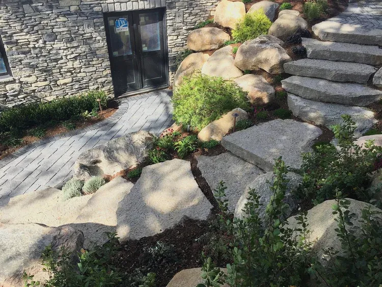 Stone garden steps and walkway beside a rock wall and black door, with shrubs and large boulders.
