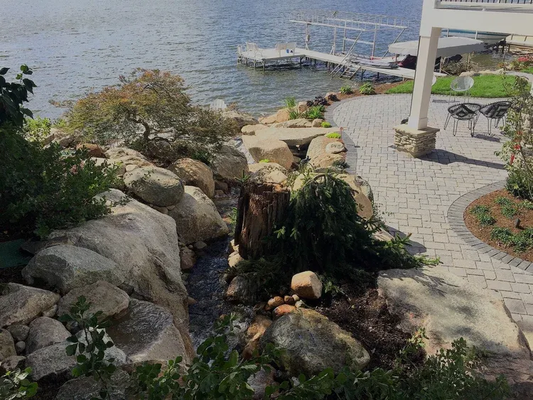 Stone path and garden beside a lakeside dock with rocks, shrubs, and patio chairs
