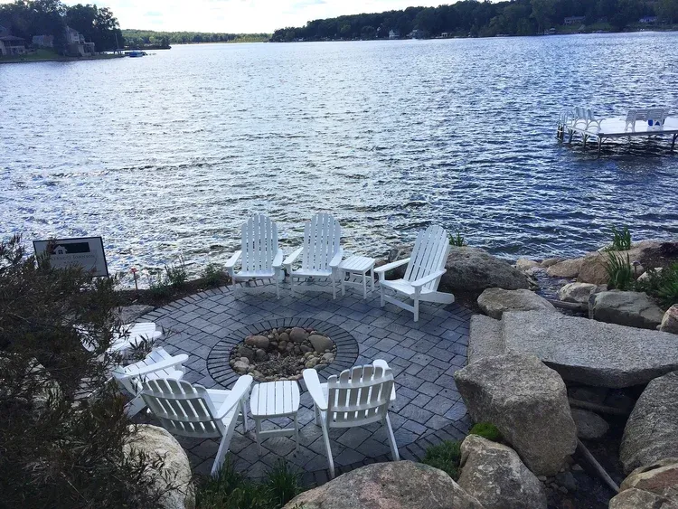 Patio chairs and table beside a rocky lakeshore with calm water and distant tree-lined shore