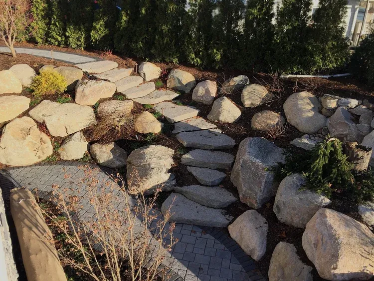 Rock garden with large boulders, stepping stones, and a narrow paved path beside shrubs and trees