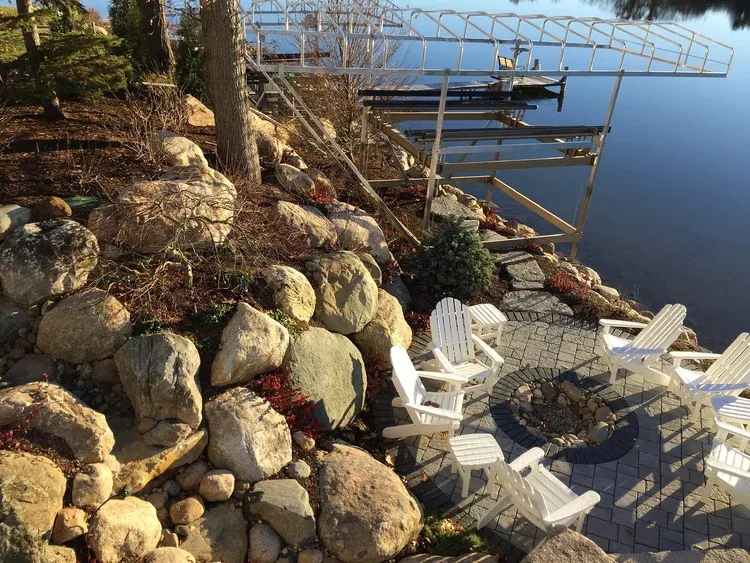 Lakeside dock with white chairs on a rocky slope beside calm blue water and a tree-lined shore