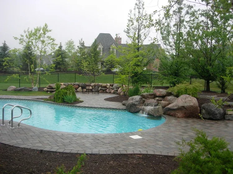 Backyard pool with stone patio, boulders, and lush green landscaping.