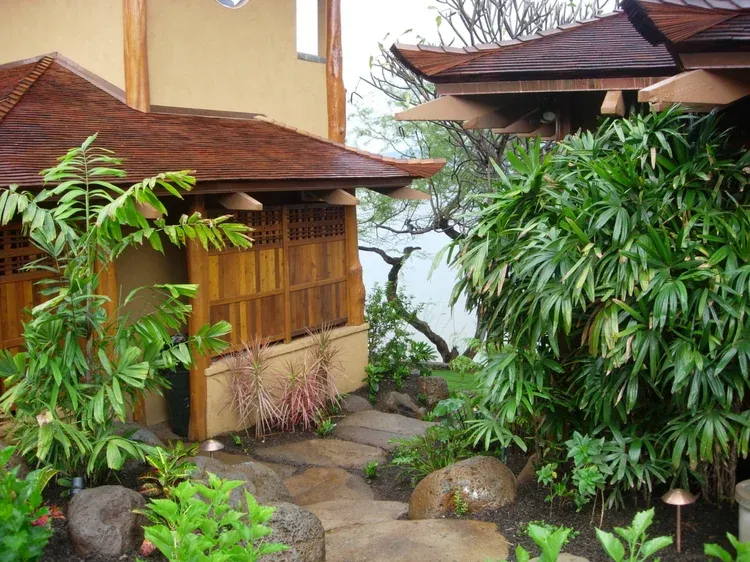 Garden courtyard with stone path, lush green plants, and a small wooden pavilion beside a house