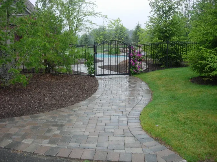 Curved stone walkway leading to a black gate in a landscaped garden with green shrubs and flowers