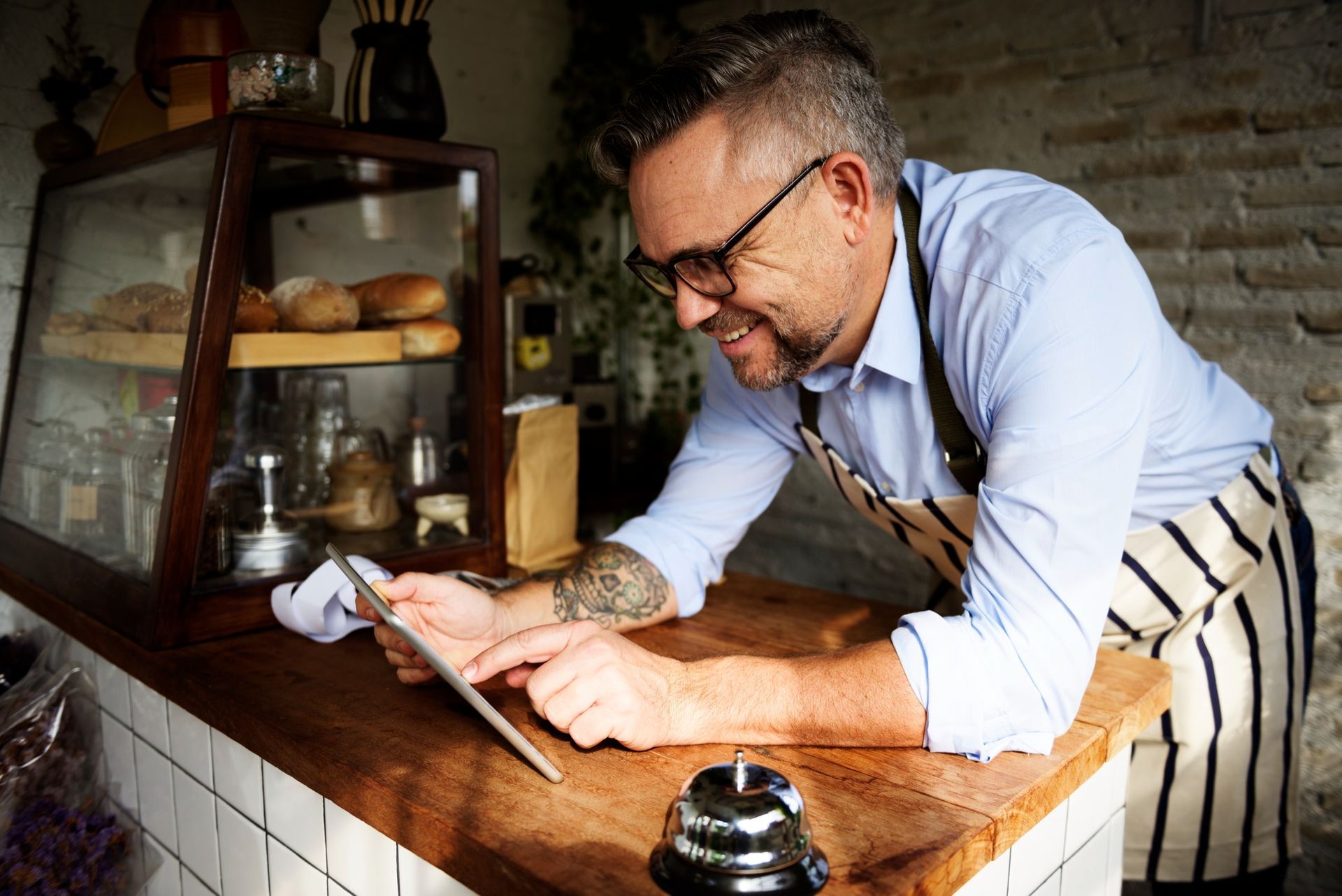 Man in apron, glasses, and tattoos smiles while using tablet at a counter in a cafe.