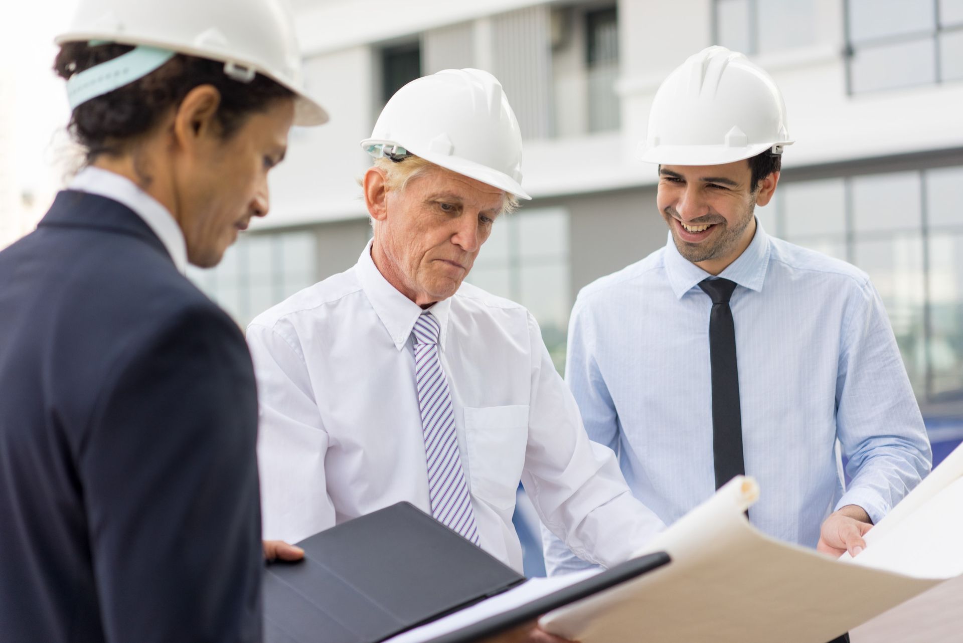 Three construction workers in hard hats examine blueprints outdoors.