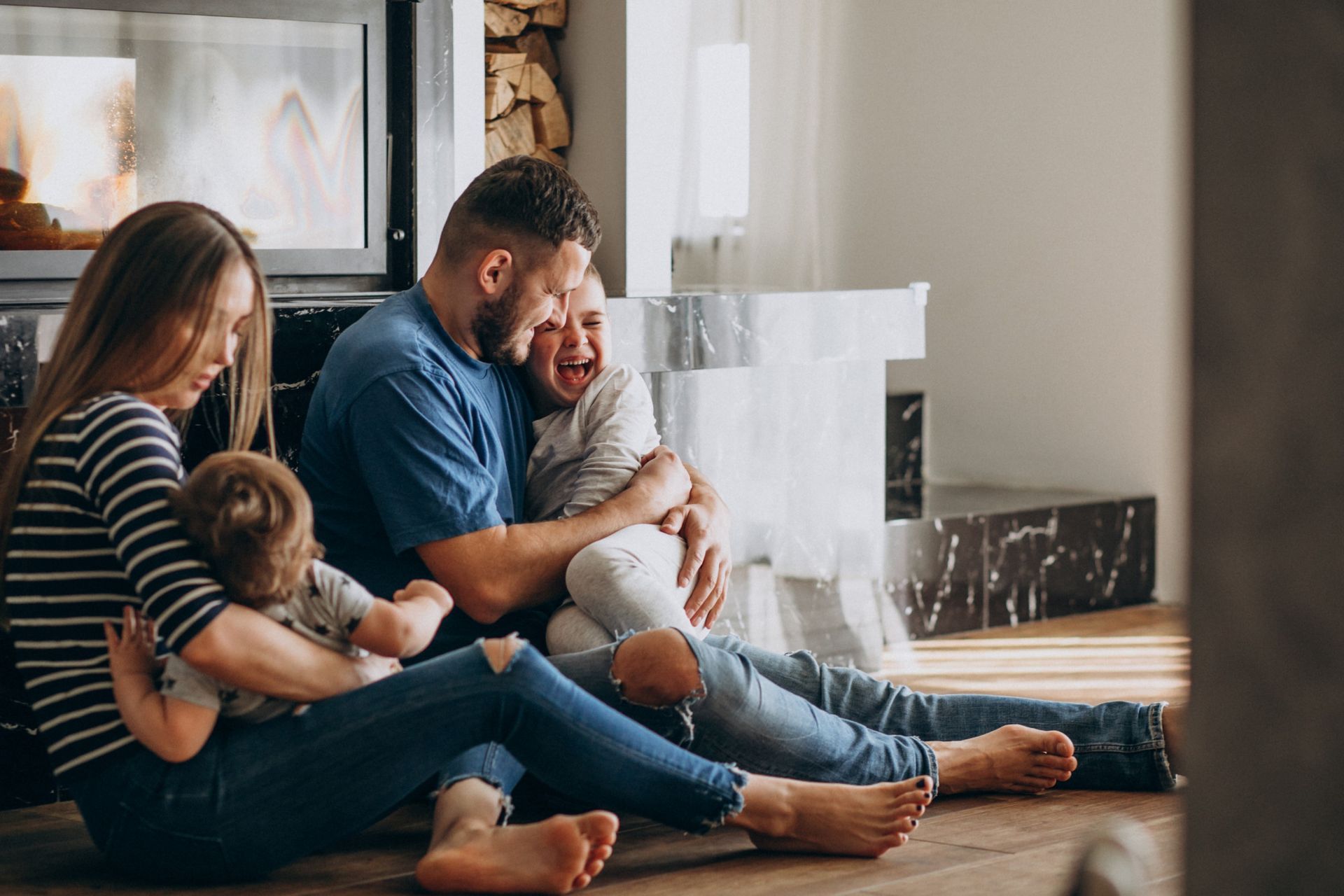 Family sitting on floor by fireplace, embracing and laughing.