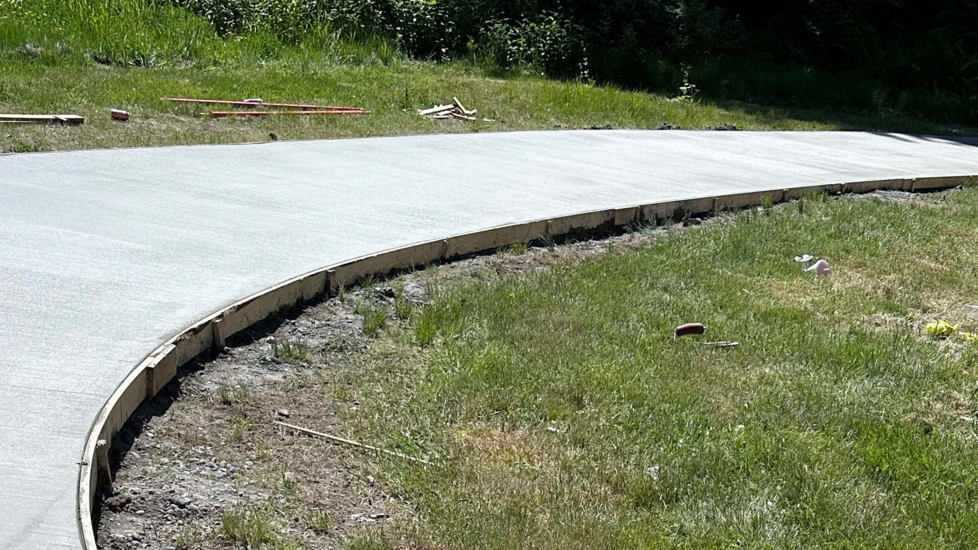 Curved concrete walkway under construction beside a grassy area with trees in the background
