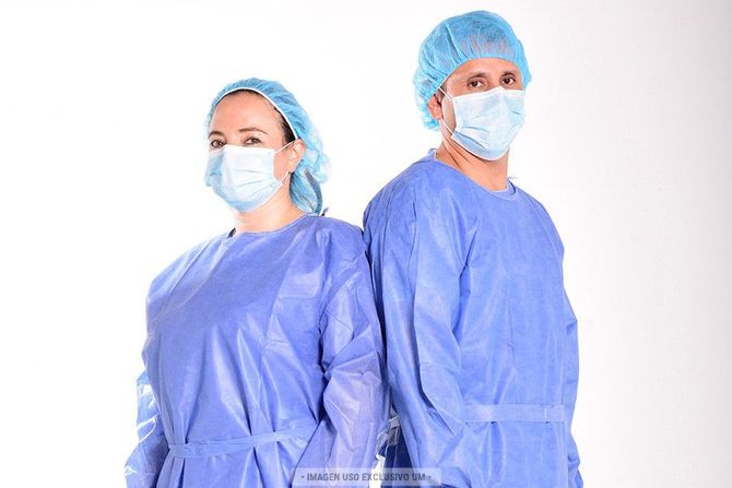 Dos trabajadores sanitarios con uniformes azules, gorros y mascarillas de pie sobre un fondo blanco.