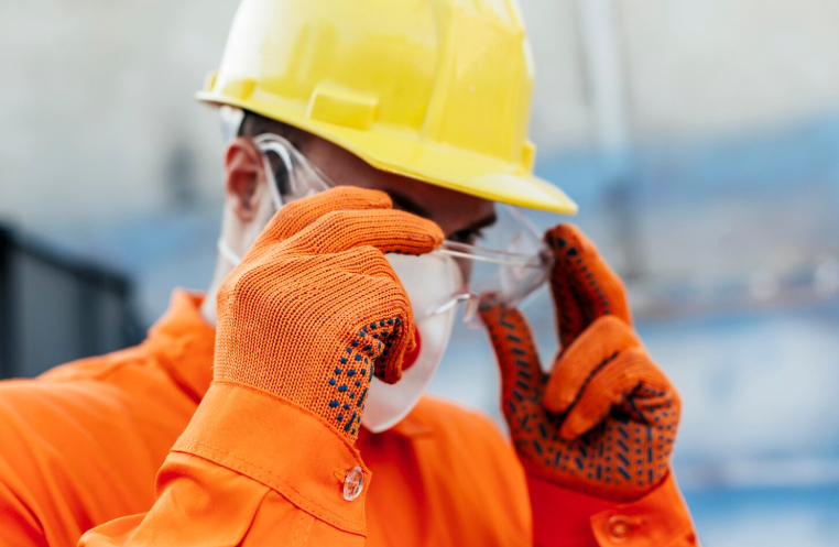 Obrero de la construcción con ropa naranja poniéndose gafas de seguridad transparentes y un casco amarillo.
