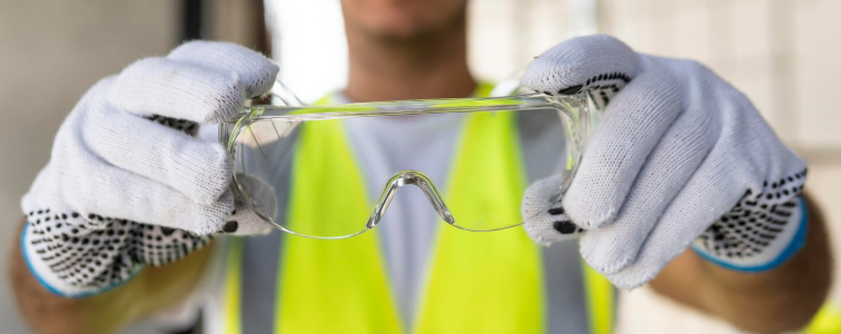 Manos con guantes blancos sosteniendo gafas de seguridad transparentes delante de un chaleco de alta visibilidad.