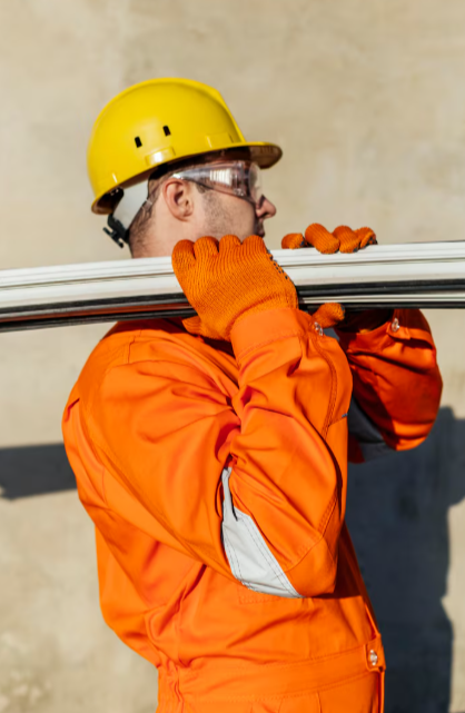 Trabajador con traje de seguridad naranja y casco amarillo cargando tuberías metálicas sobre el hombro.