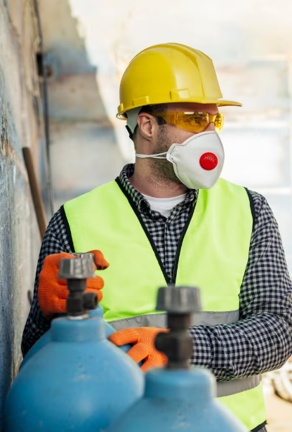 Trabajador con casco amarillo y respirador manipulando cilindros de gas en una obra.