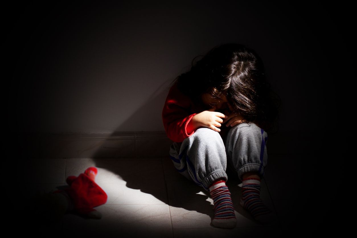 A little girl is sitting on the floor in a dark room with her head down.