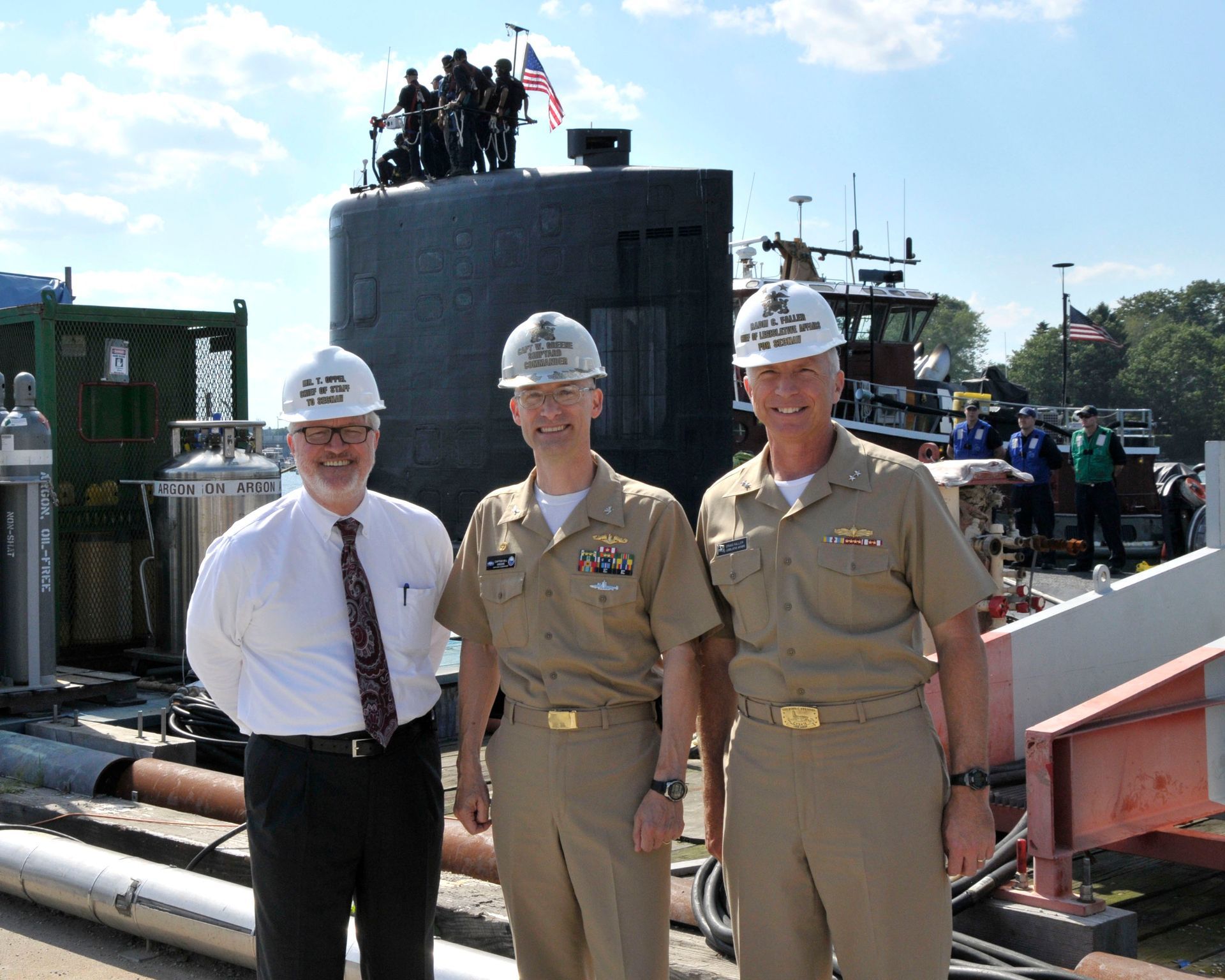 Three men in hard hats pose at a submarine exhibit outdoors on a sunny day.