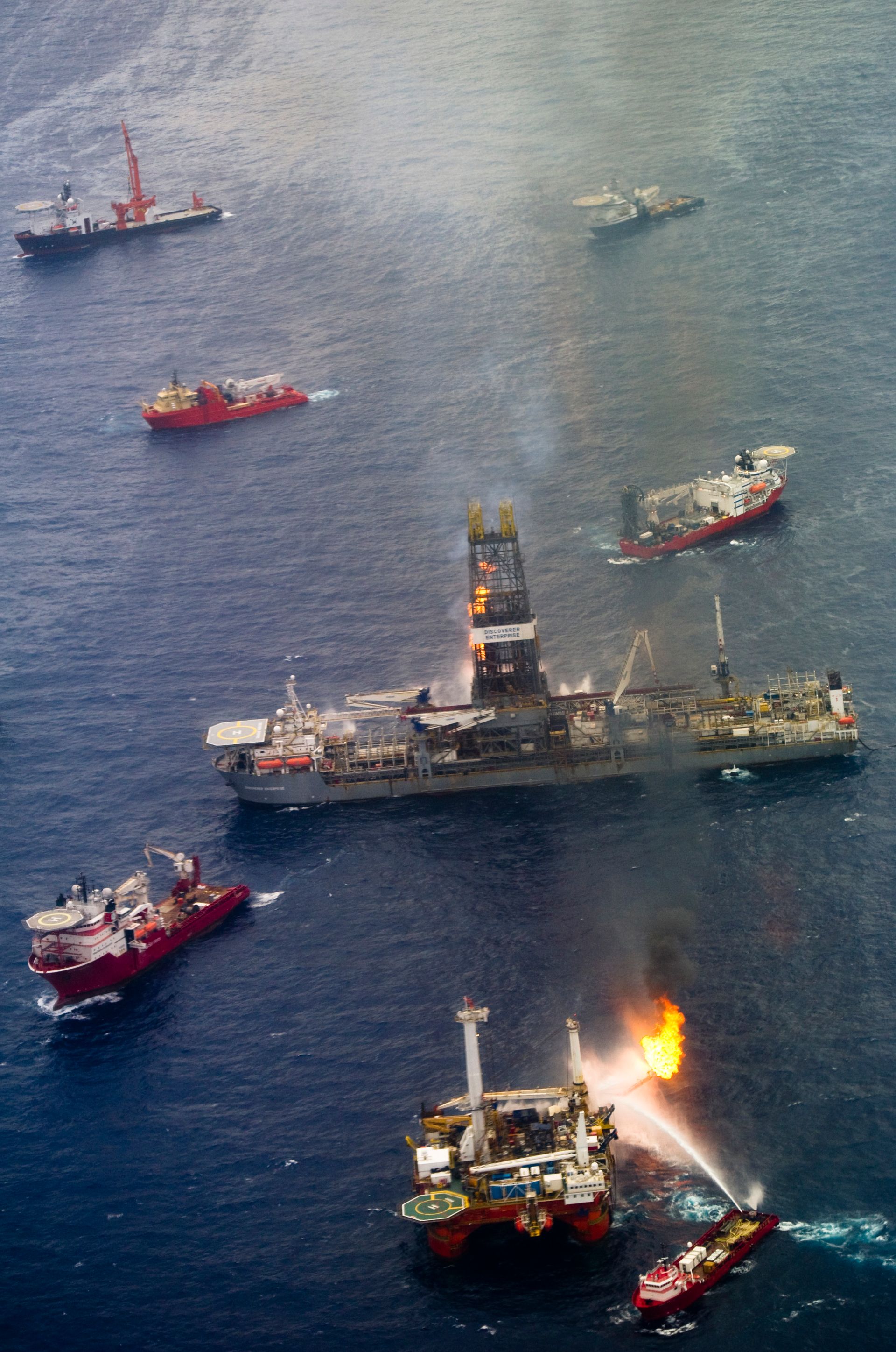 Multiple rescue boats surround a burning offshore oil rig in dark water