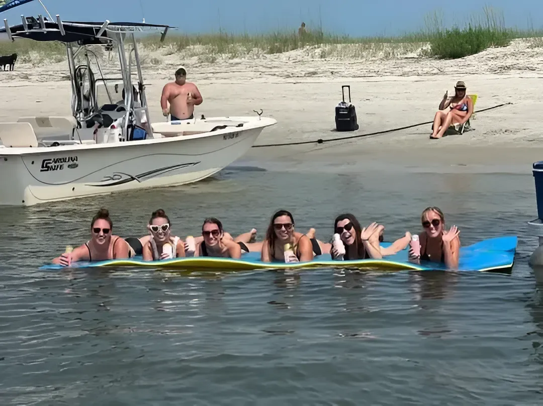 Group of people on a floating mat near a boat at a beach; others on the beach.