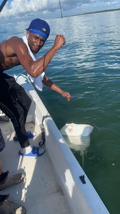 Man on a boat, reeling in a large white stingray from the water. He raises a fist, smiling. Blue sky.