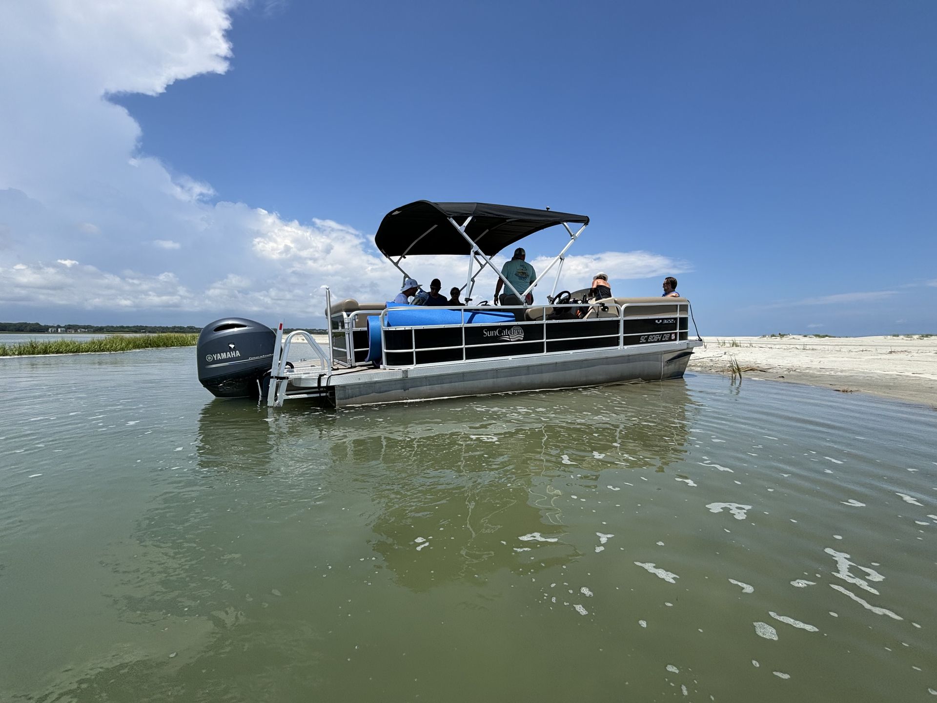 Pontoon boat cruising on dark water under a blue sky. Trees line the shore.