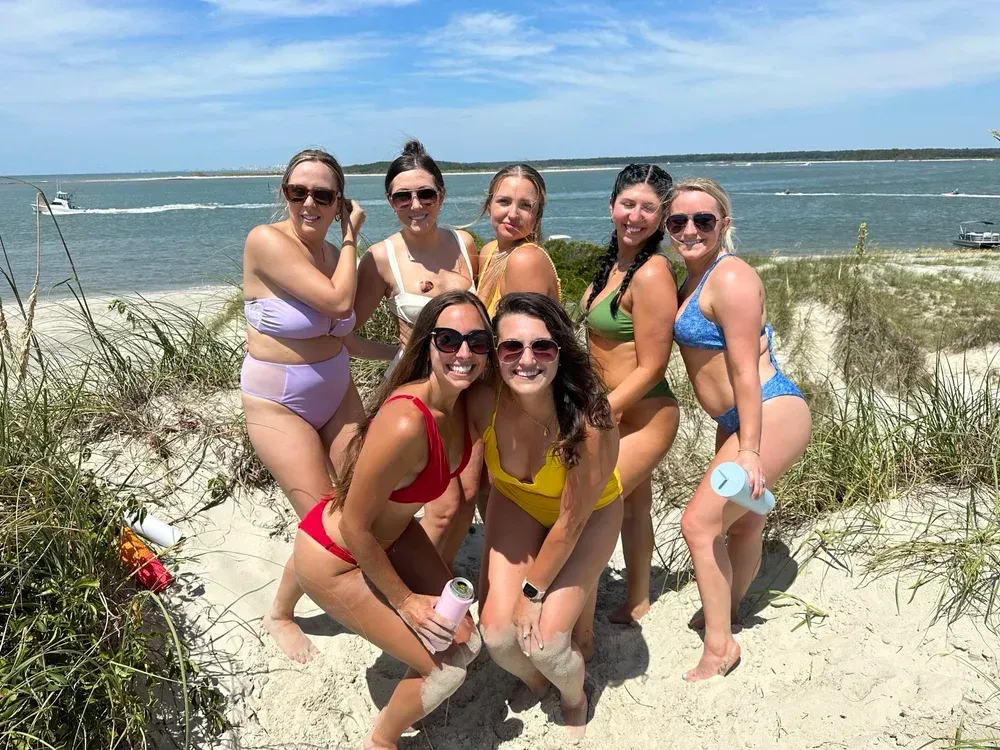 Seven women in swimsuits pose on a sandy beach, sunny day. Ocean and boats visible in background.