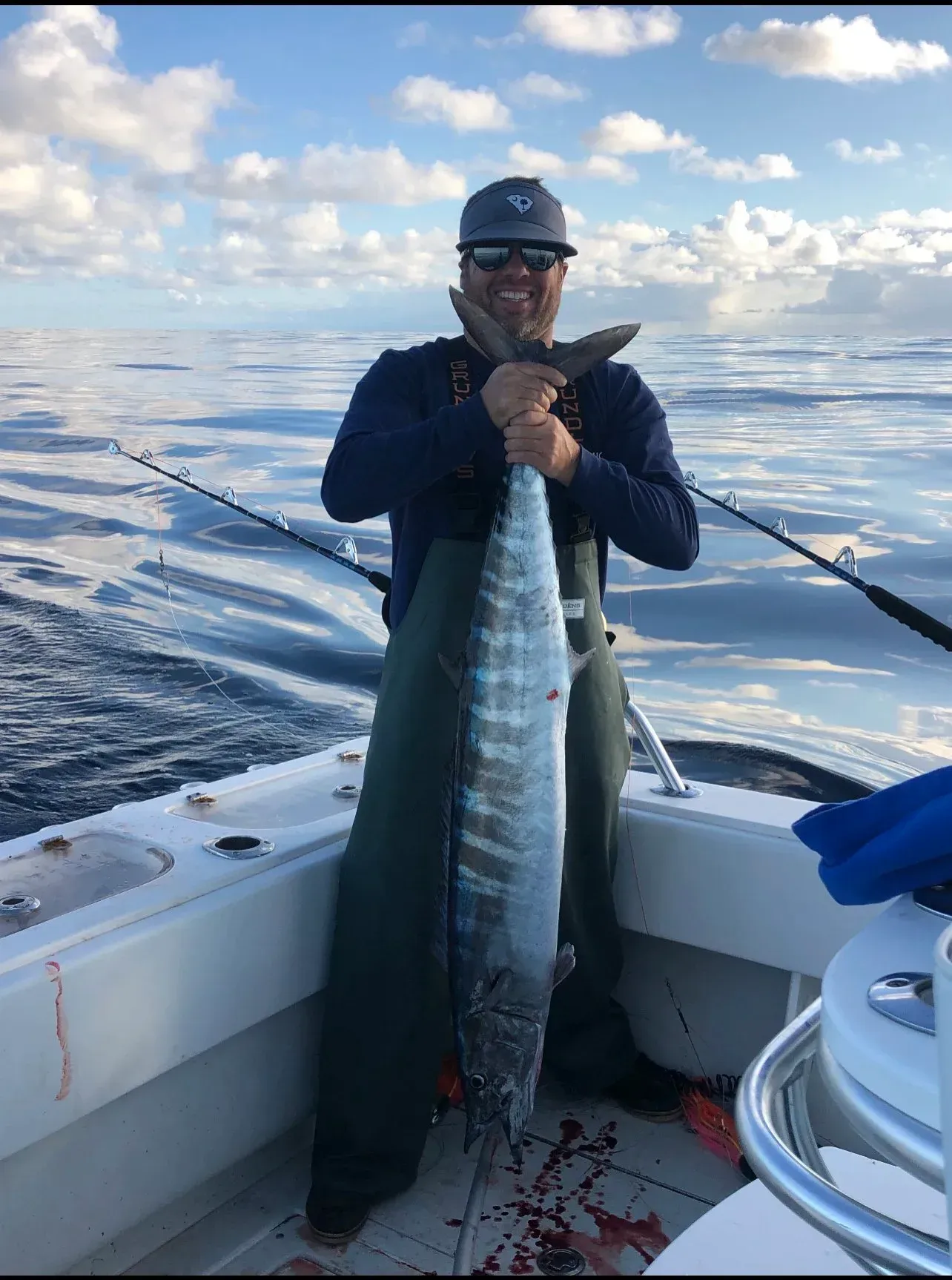 Man holding a large fish on a boat, smiling, with a blue sky background and fishing poles.