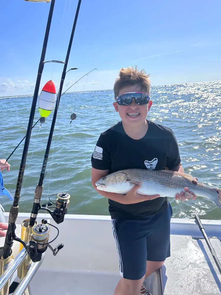 Boy on a boat smiles, holding a speckled fish. Fishing rods visible, ocean backdrop.