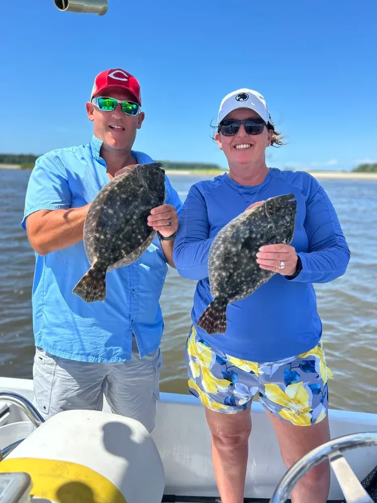 Man and woman on a boat hold up two speckled flounder, smiling. Blue sky and water are in the background.