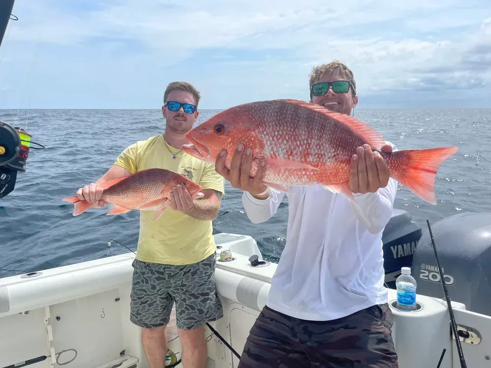 Two men on a boat hold up large red fish they caught in the ocean; blue water and sky.