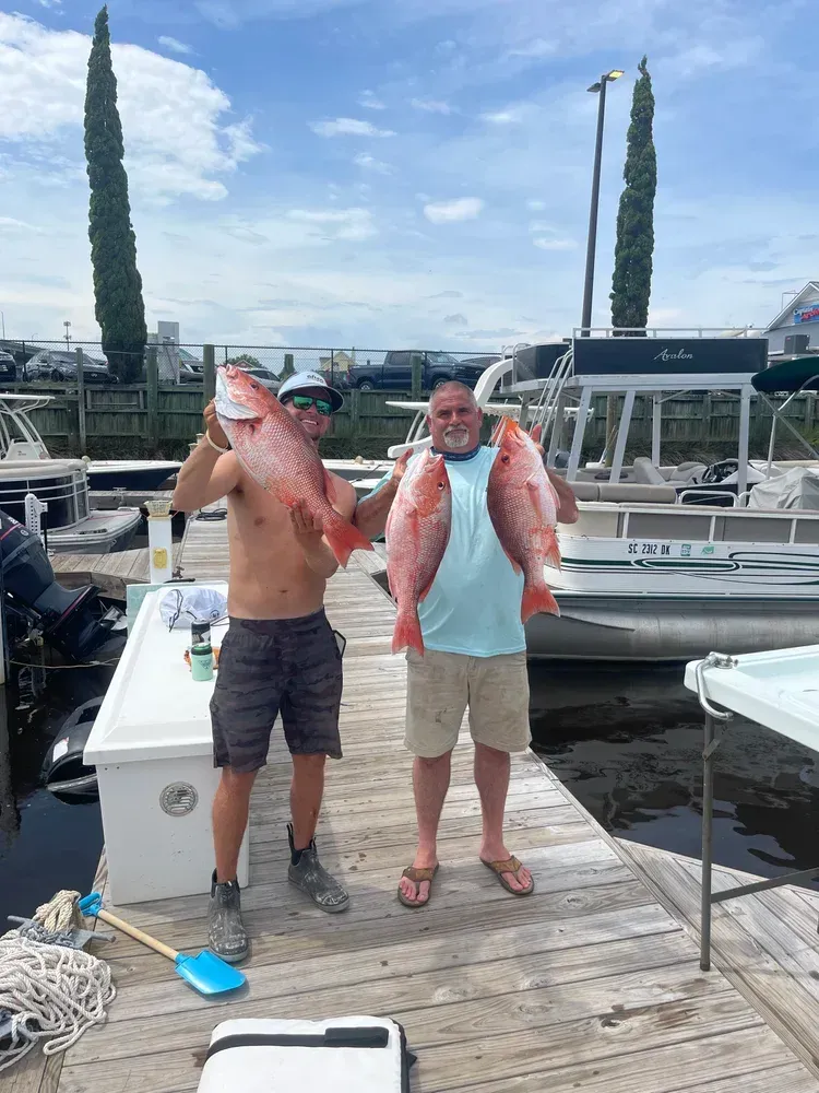 Two men holding up large red fish on a dock, boats in the background, blue sky.