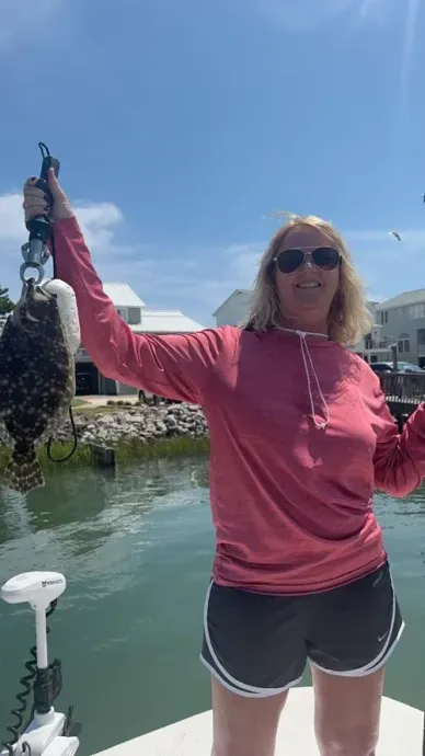 Woman holding up a fish on a scale. She is on a boat, wearing sunglasses and a pink shirt.
