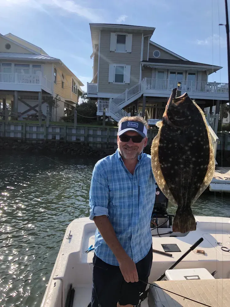 Man on a boat holding a large flounder, houses in the background. Sunny day.