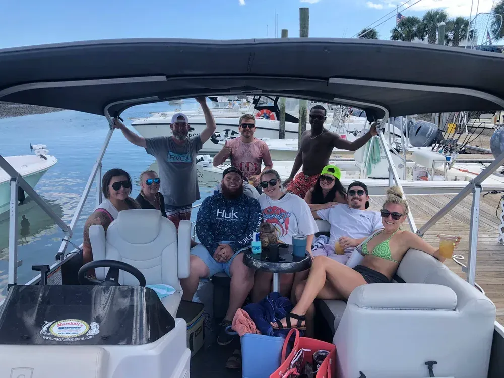 Group of people on a boat, smiling. Some have drinks. Sunny day at a marina.
