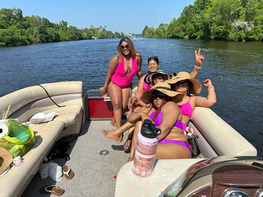 Group of people on a boat, smiling, posing in swimsuits. Surrounded by water and trees, on a sunny day.