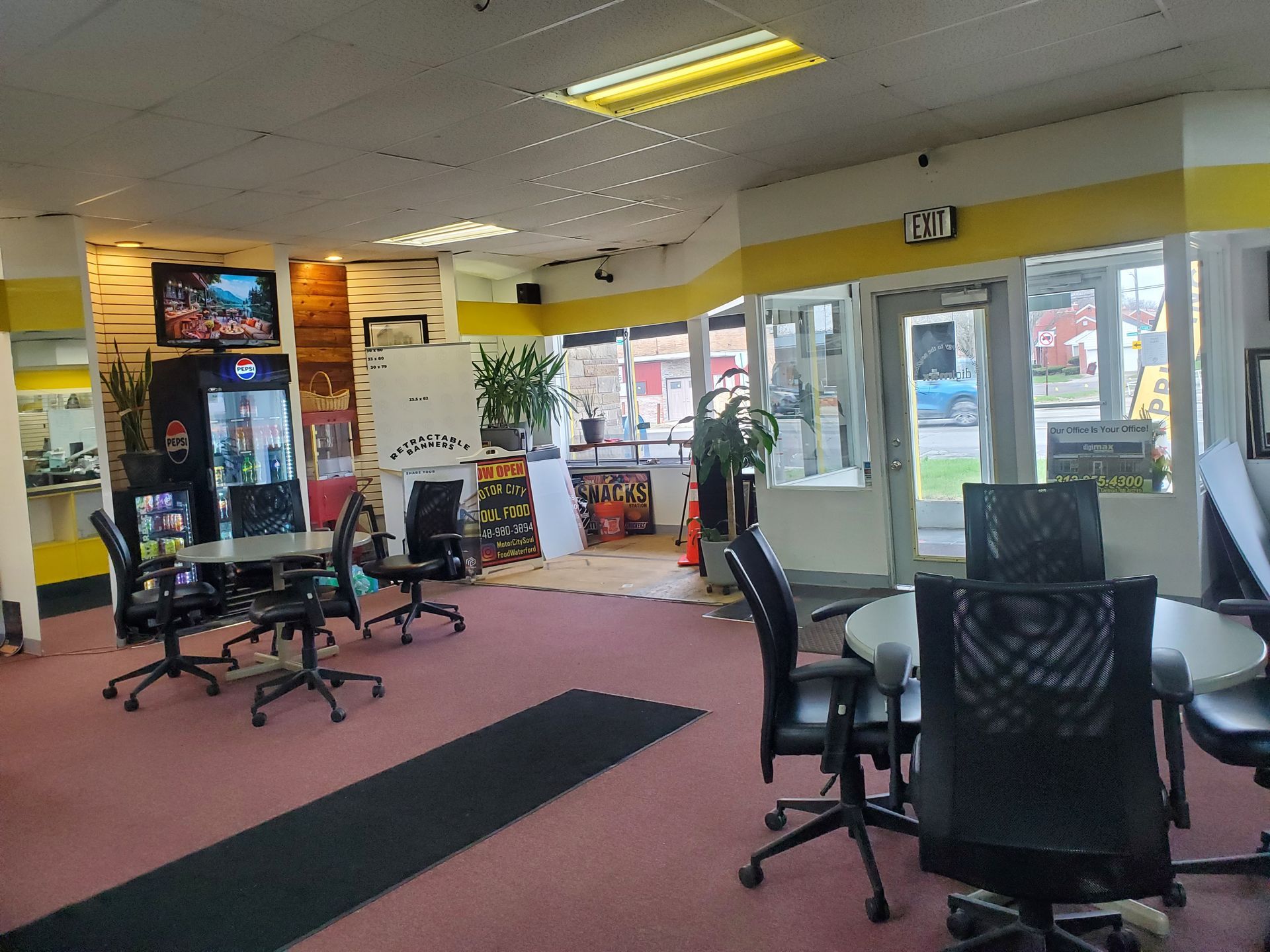 An open office space featuring desks, black rolling chairs, red carpet, and a glass-paneled entrance with yellow accents.